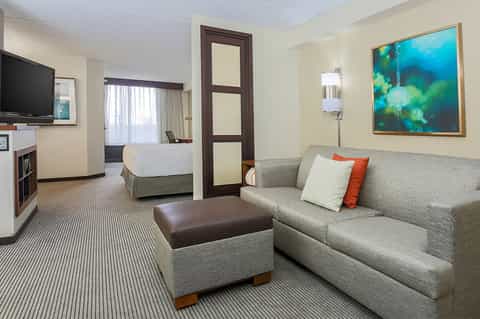 Hotel suite with queen bed, sofa, TV, and warm neutral décor with accent pillows