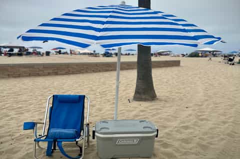 Blue and white striped beach umbrella with chair and cooler on sandy beach