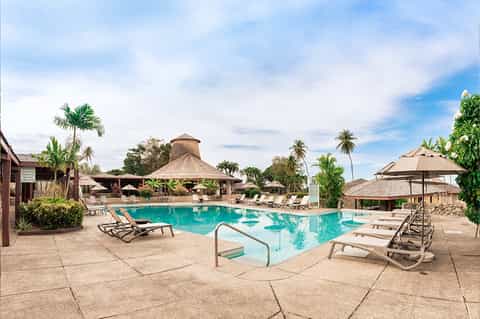 Resort pool with thatched roof structures, palm trees, and lounge chairs in tropical setting