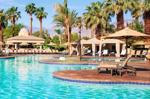 Large resort pool with lounge chairs, umbrellas, and palm trees under blue sky