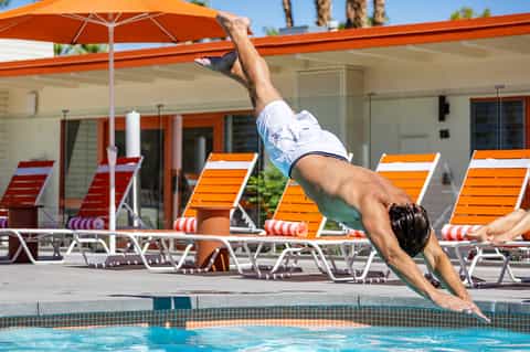 Man diving into a swimming pool with orange lounge chairs and umbrellas in the background