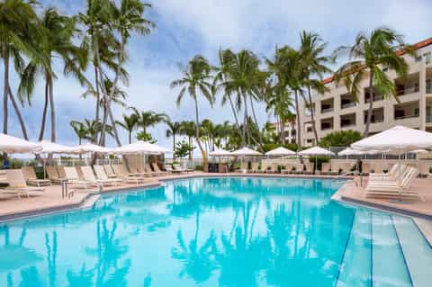 Beachfront hotel pool with palm trees, white umbrellas, lounge chairs, and multi-story building