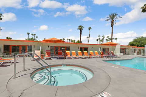 Resort pool area with hot tub, orange lounge chairs, palm trees, and mid-century modern building architecture