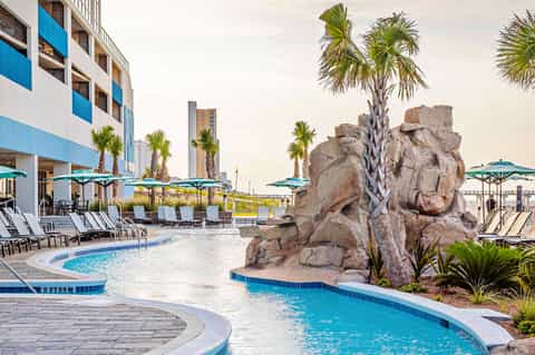 Modern resort pool area with curved pools, rock formations, palm trees, loungers, and turquoise umbrellas