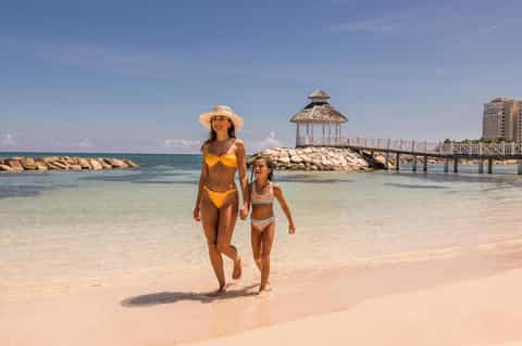 Woman and child walking on sandy beach near white gazebo and rock jetty