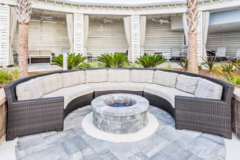 Curved wicker sectional sofa around stone fire pit on paved patio with palm trees and white pergolas