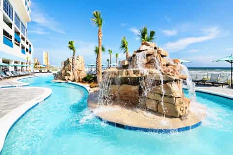 Resort outdoor pool with rock waterfall, palm trees, and beachfront hotel building under clear blue sky