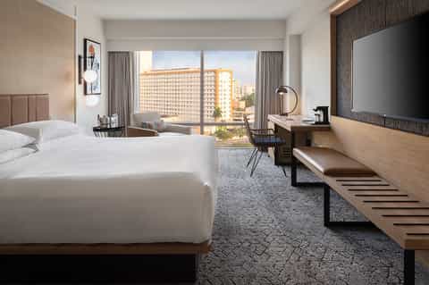 Contemporary hotel room with queen bed, city view windows, desk area, and modern furnishings overlooking urban buildings