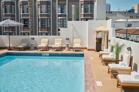Outdoor rooftop pool with lounge chairs, umbrellas, and white wall backdrop facing tall residential building