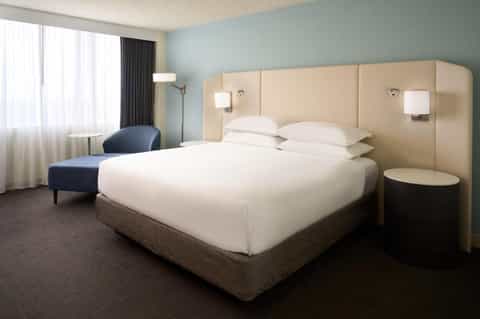 Modern hotel bedroom with queen bed, beige headboard, light blue walls, and contemporary furnishings