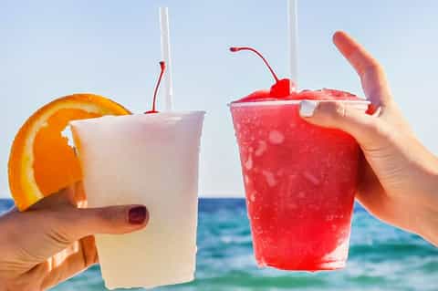 Two colorful tropical cocktails with citrus garnish held against ocean view