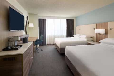 Modern hotel room with two queen beds, TV, desk, and large windows with curtains