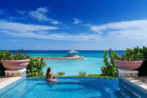 Infinity pool overlooking turquoise ocean with woman swimming, pink planters, and gazebo on distant island