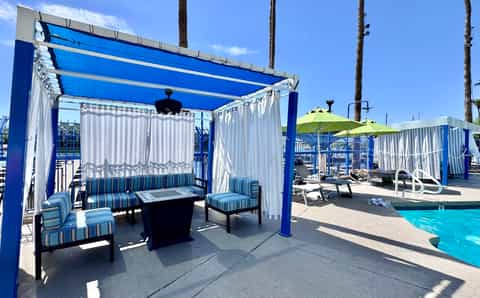 Poolside cabana with blue pergola, striped seating, and fire pit during daytime