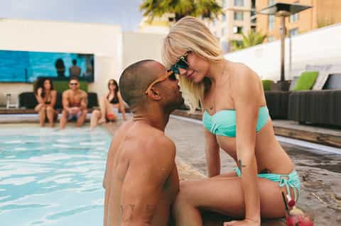 Woman in turquoise bikini interacting with man in pool at resort with other guests and palm trees visible in background