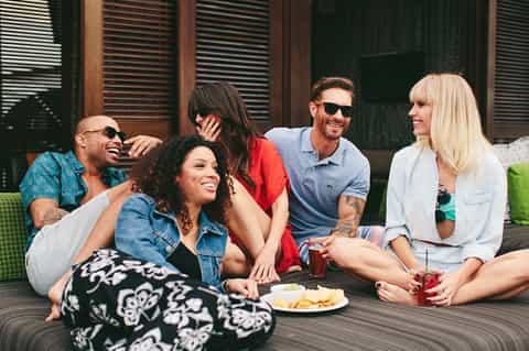 Group of six friends laughing and socializing on an outdoor deck with food and drinks