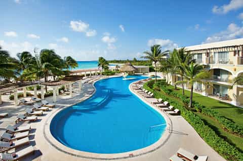 Curved blue swimming pool with palm trees, loungers, and beachfront resort building