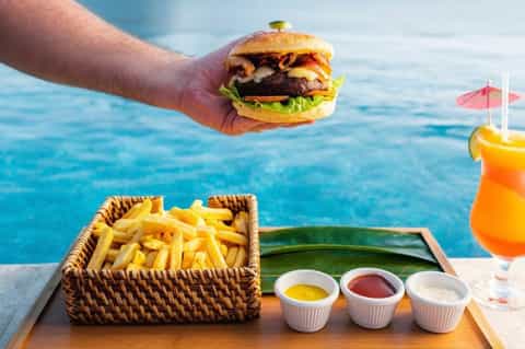 Hand holding gourmet burger with fries and tropical cocktail overlooking ocean