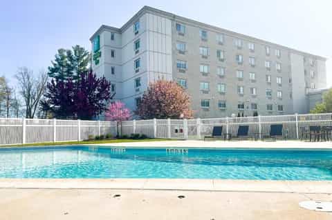 Mid-rise hotel building with swimming pool, white fence, blooming trees, and lounge chairs.