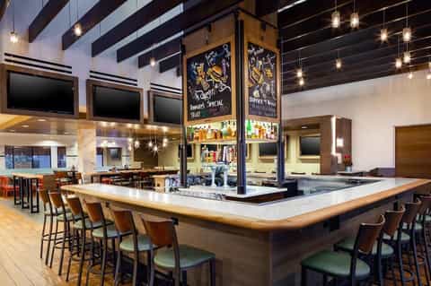 Contemporary bar and lounge with central bar, pendant lighting, dark wood beams, and multiple TV screens