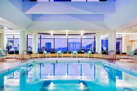 Modern indoor pool with high ceilings, floor-to-ceiling windows overlooking city skyline, and lounge seating