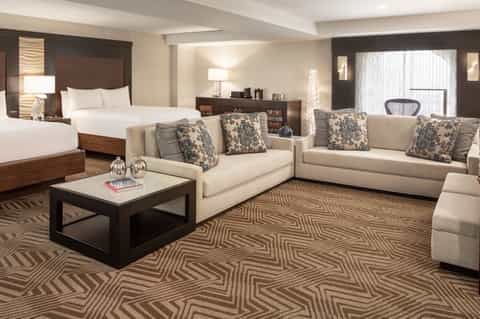 Luxury hotel suite with king bed, cream sofas, patterned pillows, and dark wood accents