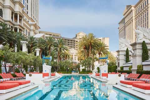Luxury resort pool flanked by red loungers with classical columns and palm trees, elegant buildings beyond
