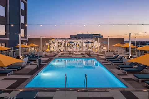 Rooftop pool at dusk with blue water, lounge chairs, yellow umbrellas, and cityscape with lit buildings