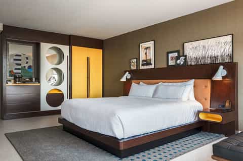 Modern hotel bedroom with wood headboard, white bedding, yellow accents, and contemporary artwork