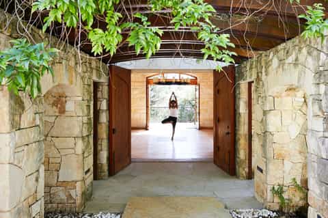 Woman in yoga pose on wooden platform visible through stone archway with green foliage overhead
