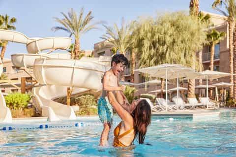 Woman and child playing in resort pool with water slide, palm trees, and white umbrellas in background