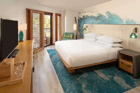 Coastal hotel bedroom with king bed, turquoise artwork, wood shutters, and balcony access