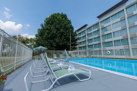 Hotel outdoor swimming pool with lounge chairs, white fence, and multi-story building backdrop