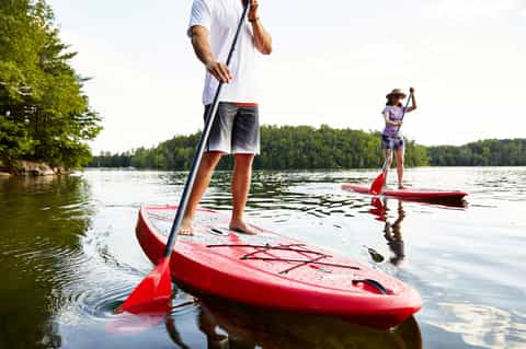 Two people paddleboarding on red kayaks in a calm lake surrounded by forested shoreline