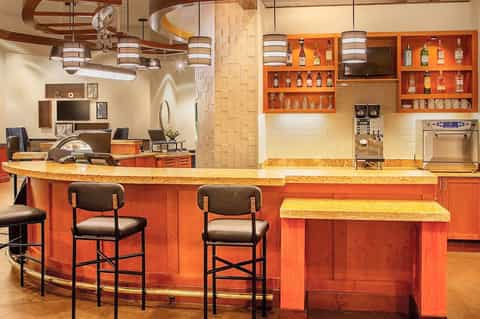Hotel bar with orange cabinetry, gold countertops, barrel chandeliers, and extensive liquor shelving