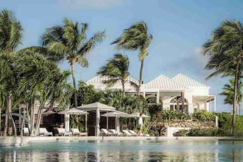 Waterfront villa with white architectural roofline, palm trees, lounge area, and pool overlooking water