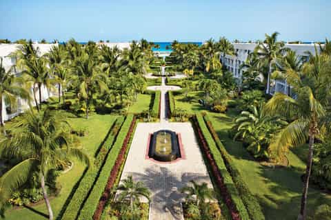 Aerial view of resort gardens with palm trees, manicured lawns, and white buildings framing a central fountain pathway