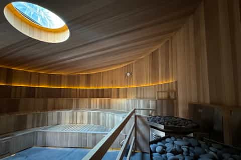 Modern sauna interior with curved wooden walls, skylight, heated benches, and stone heaters