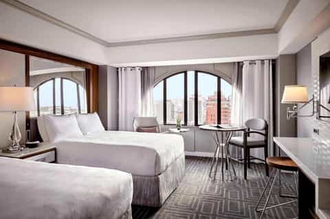 Modern hotel bedroom with two beds, city views through arched windows, and contemporary furnishings