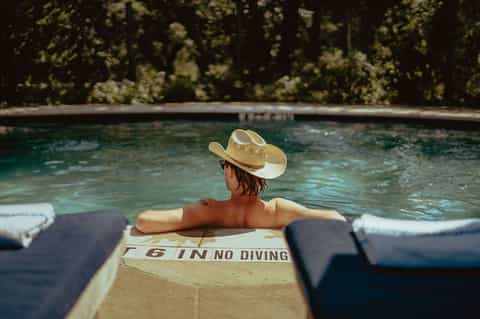 Person relaxing in resort pool wearing cowboy hat with 'No Diving' sign visible on pool deck