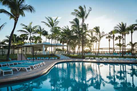 Curved resort pool surrounded by palm trees with blue lounge chairs and a poolside pavilion at dawn