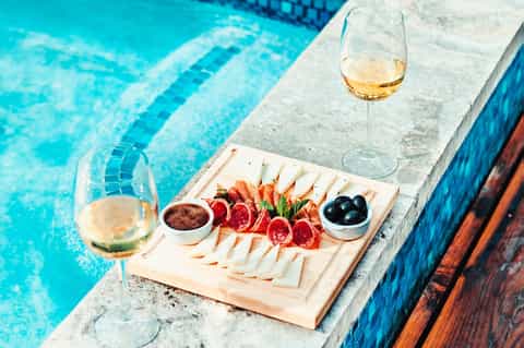 Wine and charcuterie board with cheese, cured meats, olives, and almonds beside a swimming pool