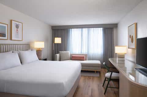 Contemporary hotel bedroom with queen bed, seating area, and large window with curtains
