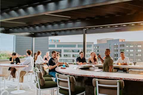 Rooftop dining area with guests at curved counter, modern black overhead structure, and city skyline views