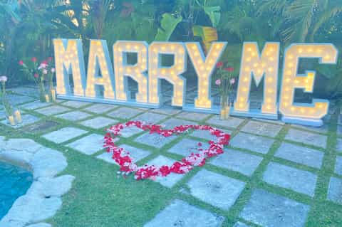 Illuminated MARRY ME sign with heart shape made of red rose petals in tropical garden setting