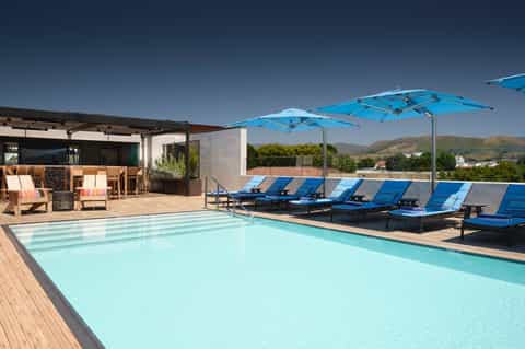 Resort rooftop pool with turquoise water, blue lounge chairs, blue umbrellas, and mountain views in background