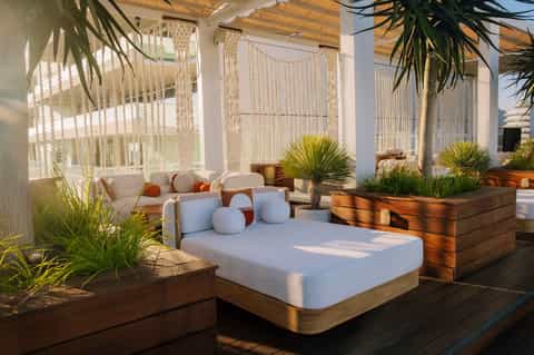 Modern covered patio lounge with white furnishings, woven pendant lights, and palm plants in sunlight