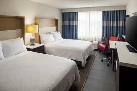 Hotel guest room with two queen beds, blue curtains, desk area, warm lighting, and contemporary furnishings