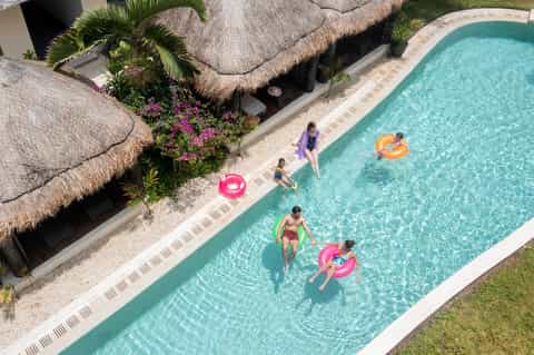 Aerial view of resort pool with families swimming and using colorful floats, thatched-roof buildings nearby