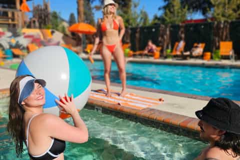 Woman holding blue and white beach ball in shallow pool with swimmers and colorful umbrellas in background
